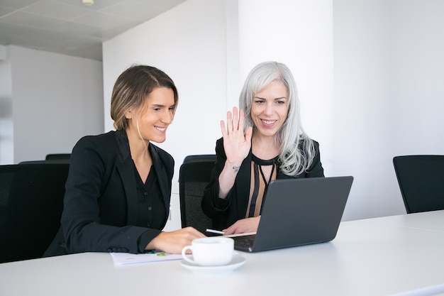 Two cheerful female colleagues using laptop for video call, sitting at table with cup of coffee, looking at display and waving hello. Online communication concept