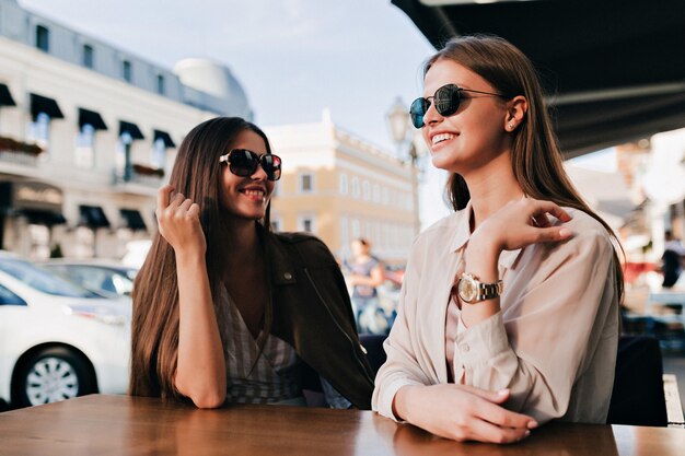 Two cheerful girls in sunglasses happily talking together with perfect smiles wearing sunglasses on square.