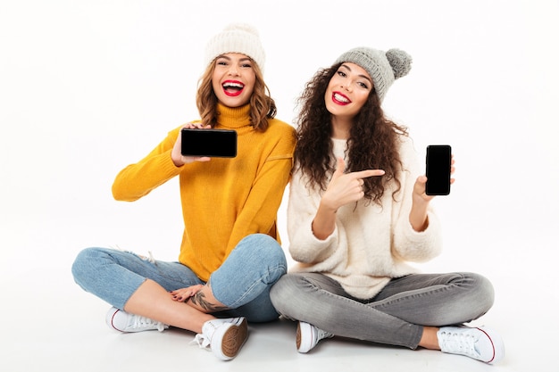 Two cheerful girls in sweaters and hats sitting on the floor together while showing blanks smartphones screens  over white wall