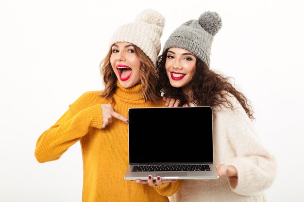 Two cheerful girls in sweaters and hats standing together while showing blank laptop computer screen  over white wall