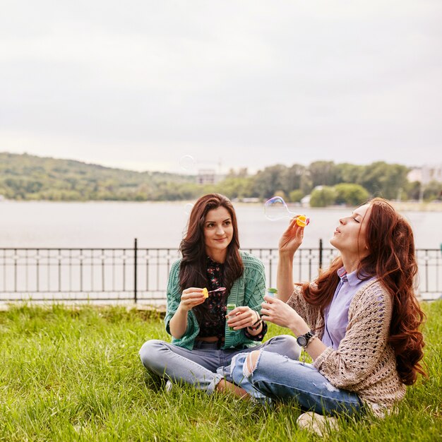 Two cheerful sisters sitting on the grass and blowing bubbles