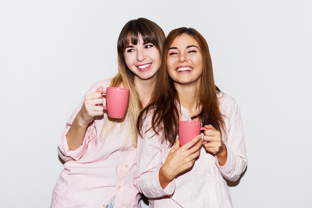 Two cheerful white  women in pink pajamas with cup of tea posing. Flash portrait.