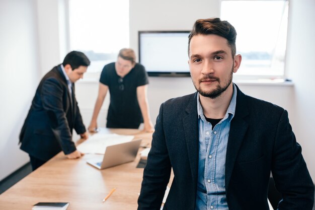 Two colleagues are working  at  table in the office. They wear black clothes. There is young guy looking to the camera on front.