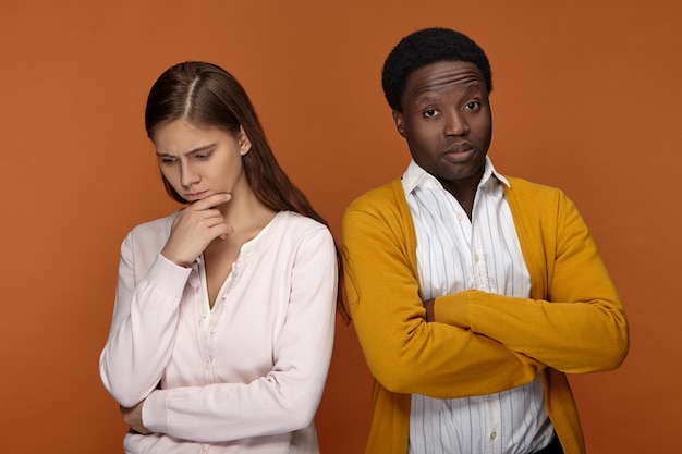 Two colleagues of different ethnicities having disagreement on business issue. African American guy with grumpy look crossing arms on his chest, not talking to worried pensive Caucasian female