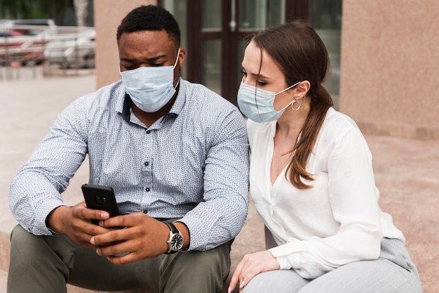 Two colleagues looking at smartphone outdoors during pandemic with face masks