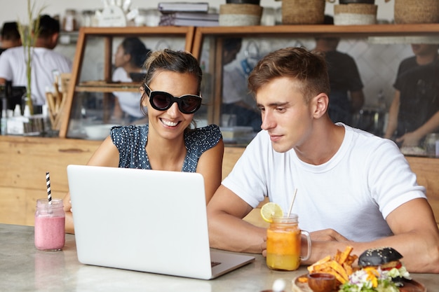 Двое коллег приятно проводят время вместе во время обеда в кафе после рабочего дня, используя портативный компьютер. Стильная женщина просматривает фотографии через социальные сети
