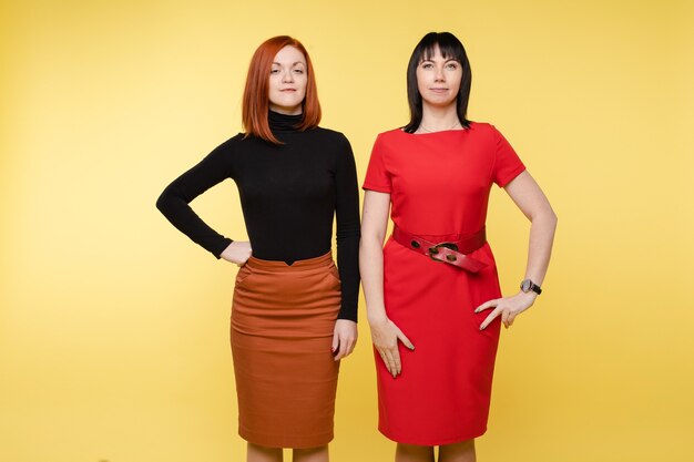 Two confident smiling business woman posing looking at camera. Fashion stylish young female standing isolated on yellow background having positive emotion