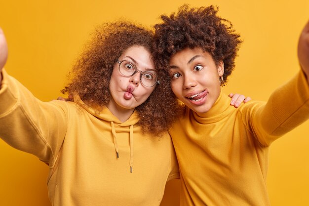 Two crazy female best friends make grimace while taking selfie embrace spreads arms forward have fun together stick out tongue make fish lips isolated over yellow wall. Mad females