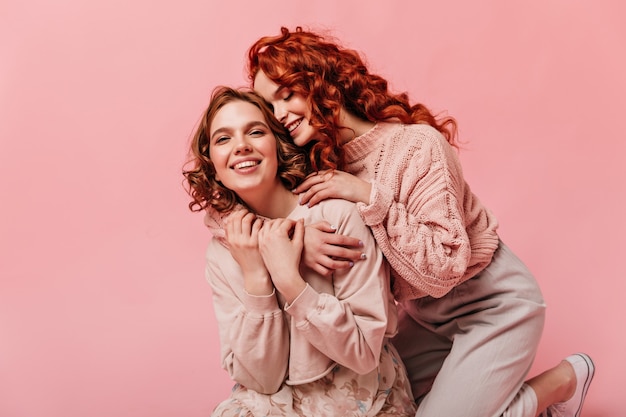 Two curly girls embracing on pink background. Blissful friends having fun together.