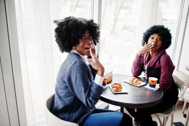 Two curly hair african american woman wear on sweaters sits at table cafe eat croissant and drink tea