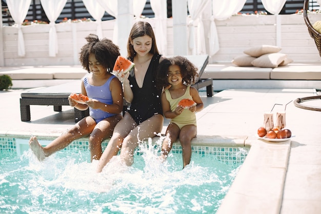 Free photo two curly multiracial sisters standing near pool stairs and their caucasian mother in the pool