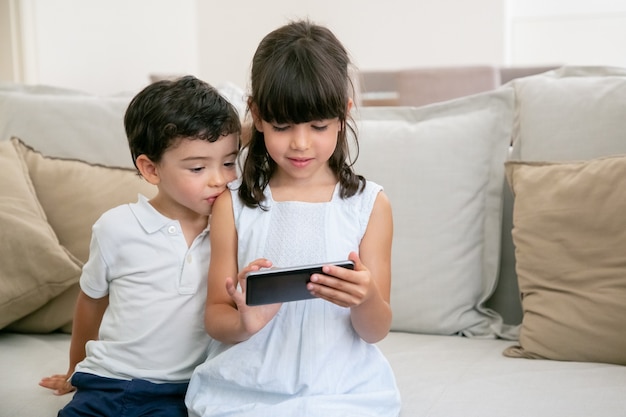 Two cute curious boy and girl sitting on couch at home and using smartphone.