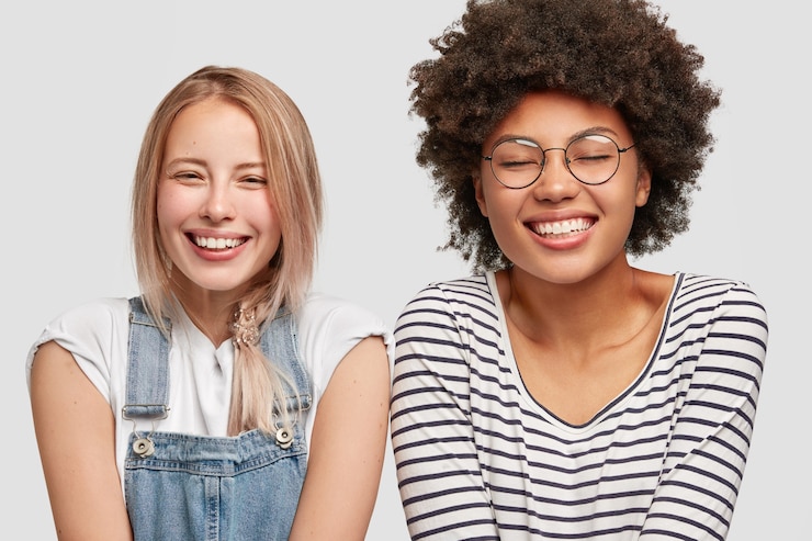 Free Photo two cute diverse women laugh positively, have broad smiles, hear funny anecdote from friend, spend free time together after having classes, express happiness, isolated over white wall