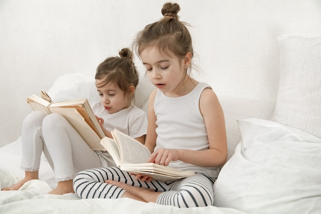 Две милые сестренки читают книгу на кровати в спальне. Понятие семейных ценностей и детской дружбы.