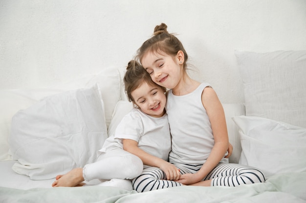 Two cute little sister girls cuddle on the bed in the bedroom.