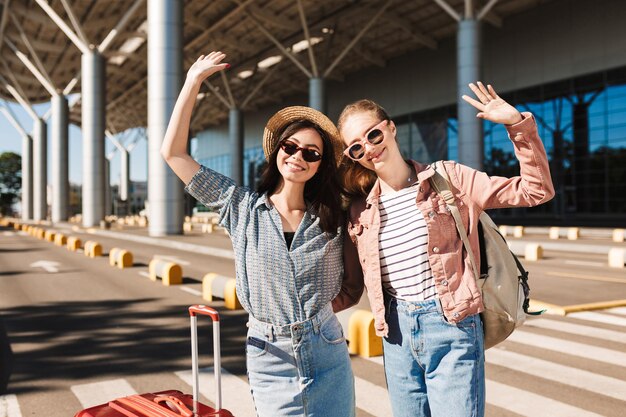 Two cute smiling girls in sunglasses happily looking in camera while raising hands up with suitcase and backpack on shoulder outdoor near airport