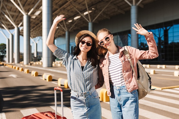 Две милые улыбающиеся девушки в солнцезащитных очках счастливо смотрят в камеру, поднимая руки вверх с чемоданом и рюкзаком на плече на открытом воздухе возле аэропорта