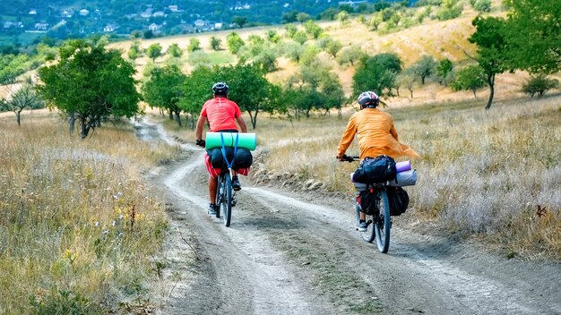 Two cyclists in helmets with bicycles full of traveler's stuff moving on the country road through rare green trees
