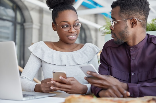 Two dark skinned female and male office managers, check notifications on mobile phones, have informal meeting, discuss something