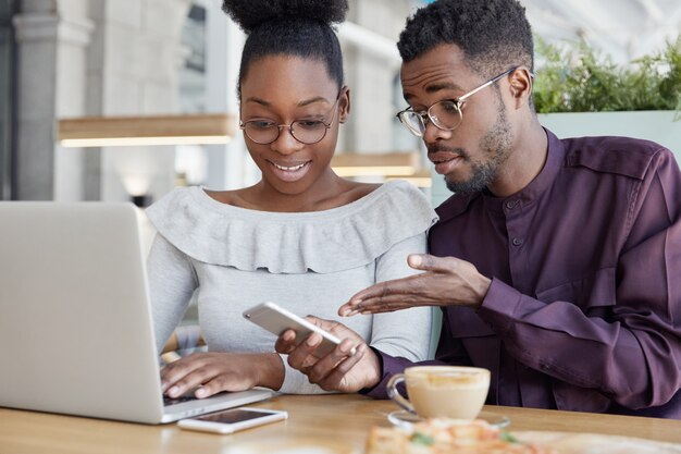 Two dark skinned male, female administrative managers check messages on cellular, keyboard on laptop computer, check information