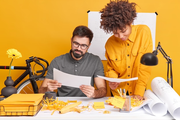 Two design workers prepare design of new building concentrated at papers pose at desktop try to find right solution 