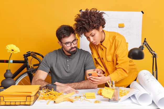 Two diverse members of working team think over cretive ideas for future project woman shows design plan in smartphone on webpage pose at desktop 