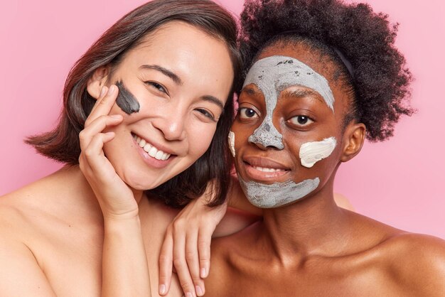 Две разные женщины наносят глиняную маску на лицо, улыбаются, с радостью стоят без рубашки в помещении, имеют дружеские отношения, имеют хорошее состояние кожи, изолированные на розовом фоне.