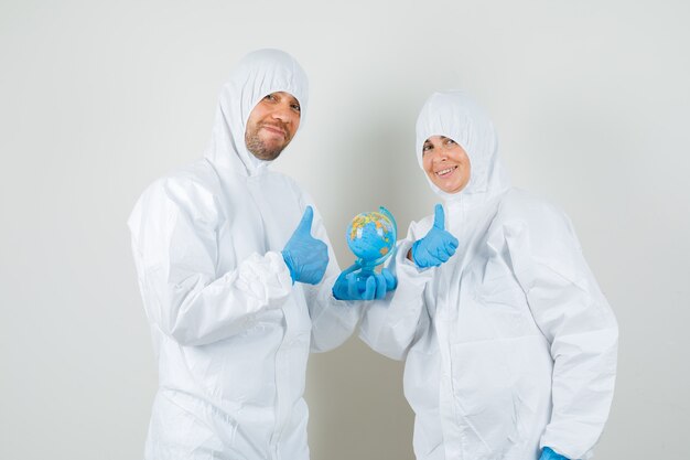 Два врача держат земной шар, показывая пальцы вверх в защитных костюмах