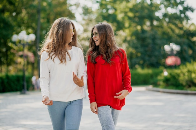 Two elegant girls with wavy long hairstyles are wearing bright shirts are talking and walking around the city in warm sunny day