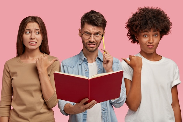 two embarrassed women of different race point at puzzled guy, suggest to ask him this question as they do not know answer, stand together against pink wall. Educational theme