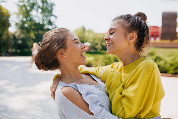 노란색과 파란색 스웨터를 입은 두 명의 감정적인 여자 친구가 공원을 걷는 동안 웃고 껴안고 있습니다