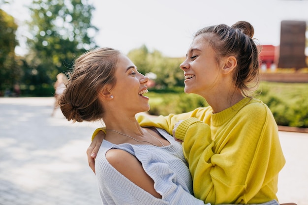 Two emotional female friends wearing in yellow and blue sweaters is laughing and hugging while walking in the park