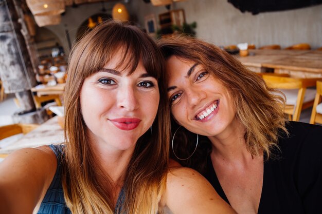 Две европейские подруги-кавказцы с естественным макияжем и короткими волосами делают селфи в летнем кафе