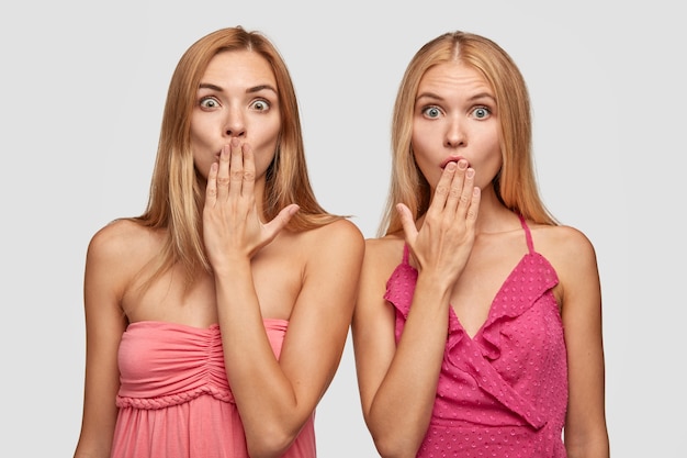 Two European young women has long light hair, cover mouth with palm, state with bugged eyes