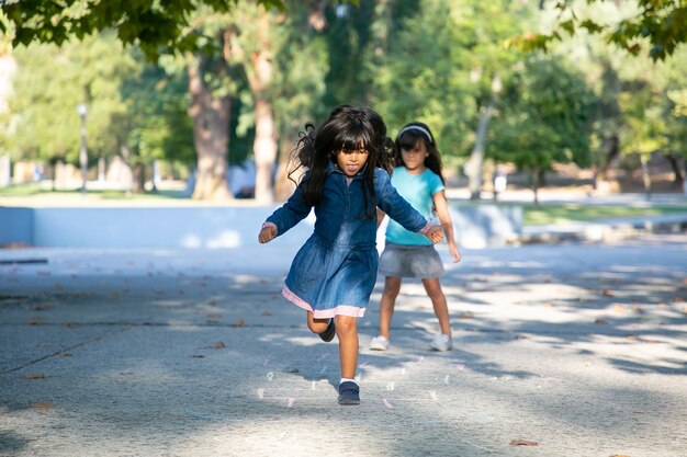 Две возбужденные черноволосые маленькие девочки играют в классики в городском парке. Полная длина, копия пространства. Концепция детства