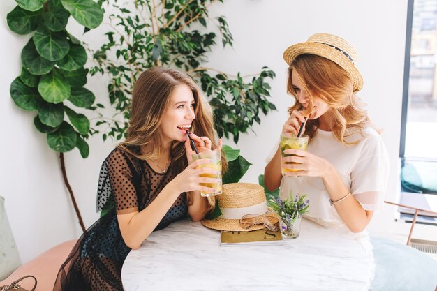 Two excited girls sitting at the table with straw hat on it and talking about something funny