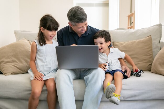 Two excited happy kids watching content on laptop with their dad while sitting on couch at home