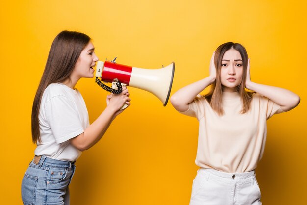 黄色の壁に隔離されたポーズのカジュアルなTシャツデニムの服を着た2人の興奮した若い女性のガールフレンド。人々のライフスタイルの概念。コピースペースをモックアップします。メガホンで叫び、手を広げます