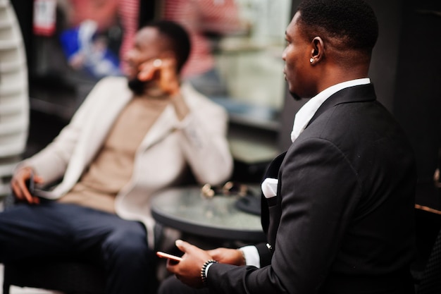 Free photo two fashion black men sitting outdoor with mobile phone fashionable portrait of african american male models wear suit coat and hat