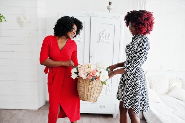 Две модные африканские американки в вечернем платье стоят с корзиной цветов на руках на фоне старого винтажного гардероба в белой комнате