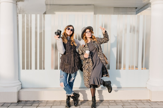 Two fashionable joyful smiling women having fun on sunny street in city. Stylish look, travelling together, wearing modern trend clothes, walking with coffe to go, expressing positive emotions.