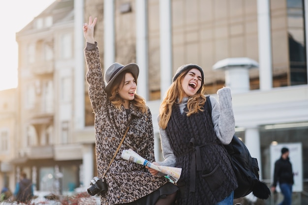 Two fashionable joyful smiling women jumping over city. Stylish look, travelling together, wearing modern trend clothes, walking with coffe to go, expressing positive emotions.