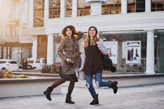 Two fashionable joyful smiling women jumping over city. Stylish look, travelling together, wearing modern trend clothes, walking with coffe to go, expressing positive emotions.