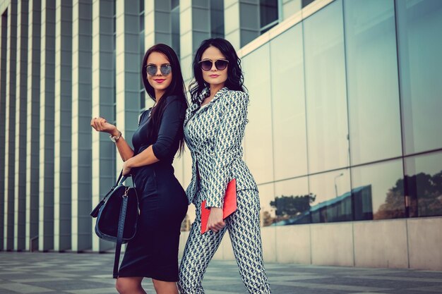 Two fashionable women in stylish clothes and sunglasses posing in a middle of business urban district.