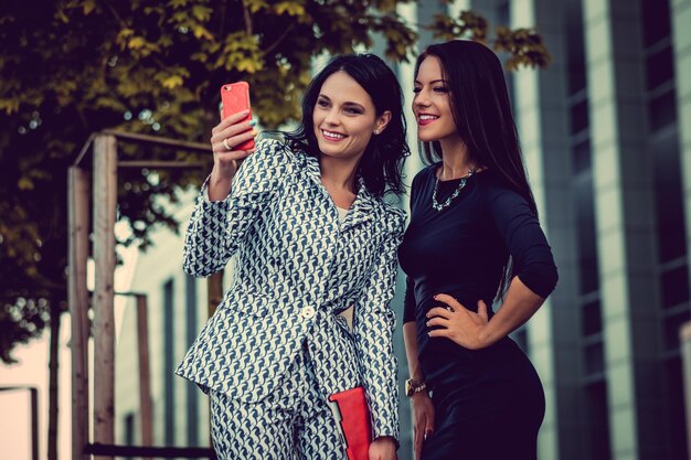 Two fashionable women in stylish clothes and sunglasses posing in a middle of business urban district.