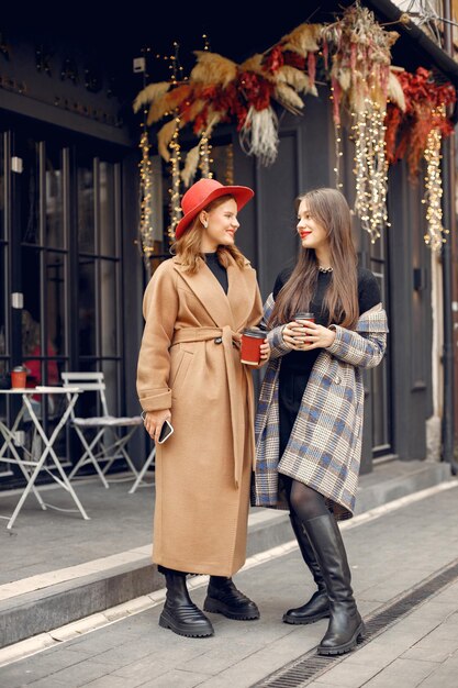 Two female best friends spending time together. Two young girls standing on the terrace in a cafe holding a coffee. Brunette girl has red lips, red head girl wearing brown coat and red hat.