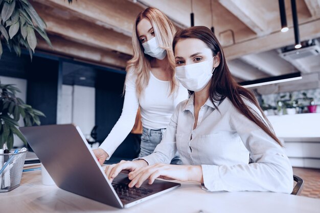 Две женщины-коллеги, работающие в офисе вместе в медицинских масках