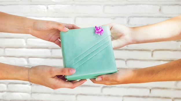 Free photo two female friend's hand holding green gift box in front of brick wall