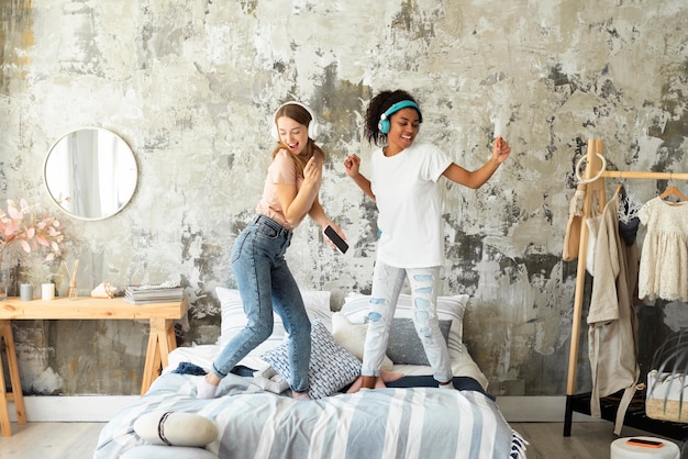 Two female friends dancing together on bed while listening to music