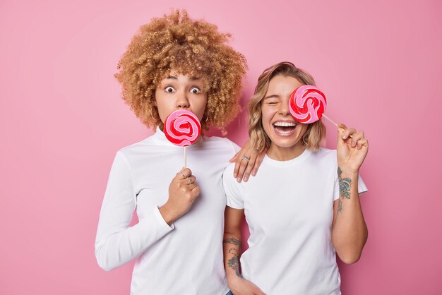 Two female friends hold big caramel candies on sticks have fun sweet tooth dressed in casual clothes stand next to each other isolated over pink background Friendship and confectionery concept